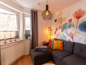 A modern living room with a sectional sofa, a radiator, a window with blinds, a hanging light fixture, and a floral wall mural. - Holiday Apartment With 2 Bedrooms (Braunlage)