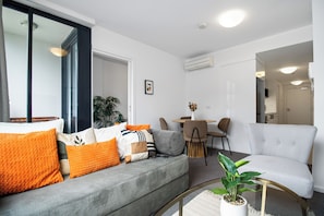 A modern living room with a grey sofa, orange cushions, a white armchair, and a coffee table with a plant. - On the Boulevard - Styled 1BR with Views (Melbourne)