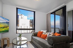 A modern living room with a large window, a sofa with orange cushions, a small round table with a plant, and a painting on the wall. - On the Boulevard - Styled 1BR with Views (Melbourne)