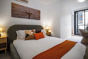 Un dormitorio con cama, almohadas, una mesita de noche con lámpara, aire acondicionado en la pared y ventana con vistas.
