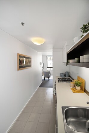 A modern kitchen with a stainless steel sink, a gas stove, and a countertop with a toaster. There is a chair in the background and a painting on the wall. - On the Boulevard - Styled 1BR with Views (Melbourne)