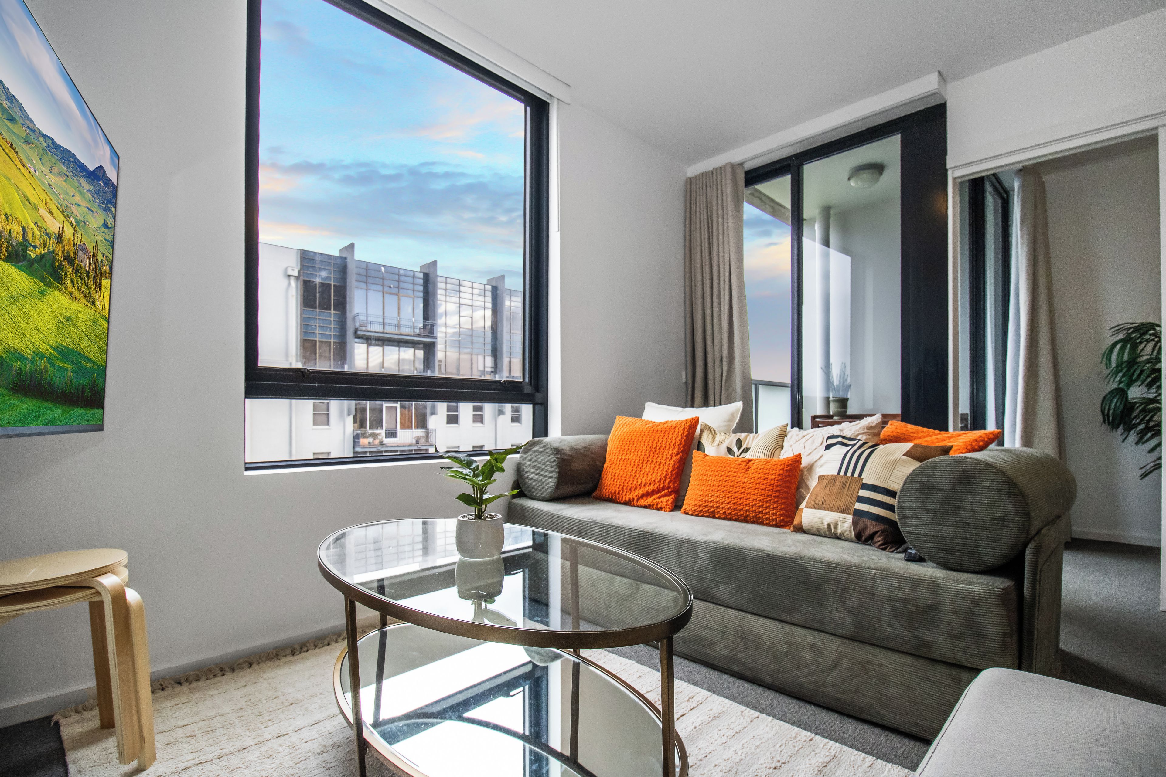 Un salon moderne avec une table basse en verre, un canapé agrémenté de coussins orange et une grande fenêtre donnant sur la ville.