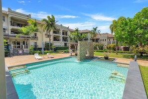 A swimming pool with lounge chairs, surrounded by a building with balconies and palm trees. - Belle Escapes Cairns One Condo with Private Pool (Cairns)