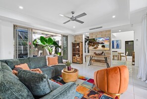 A modern living room with a sofa, armchair, and a coffee table. There is a ceiling fan, a television, and a bookshelf. - Belle Escapes Cairns One Condo with Private Pool (Cairns)