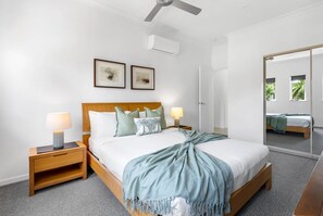Ein Hotelzimmer mit Bett, Nachttischen, Deckenventilator, Wandklimaanlage, gerahmten Kunstwerken und einem Spiegel, der den Raum widerspiegelt.