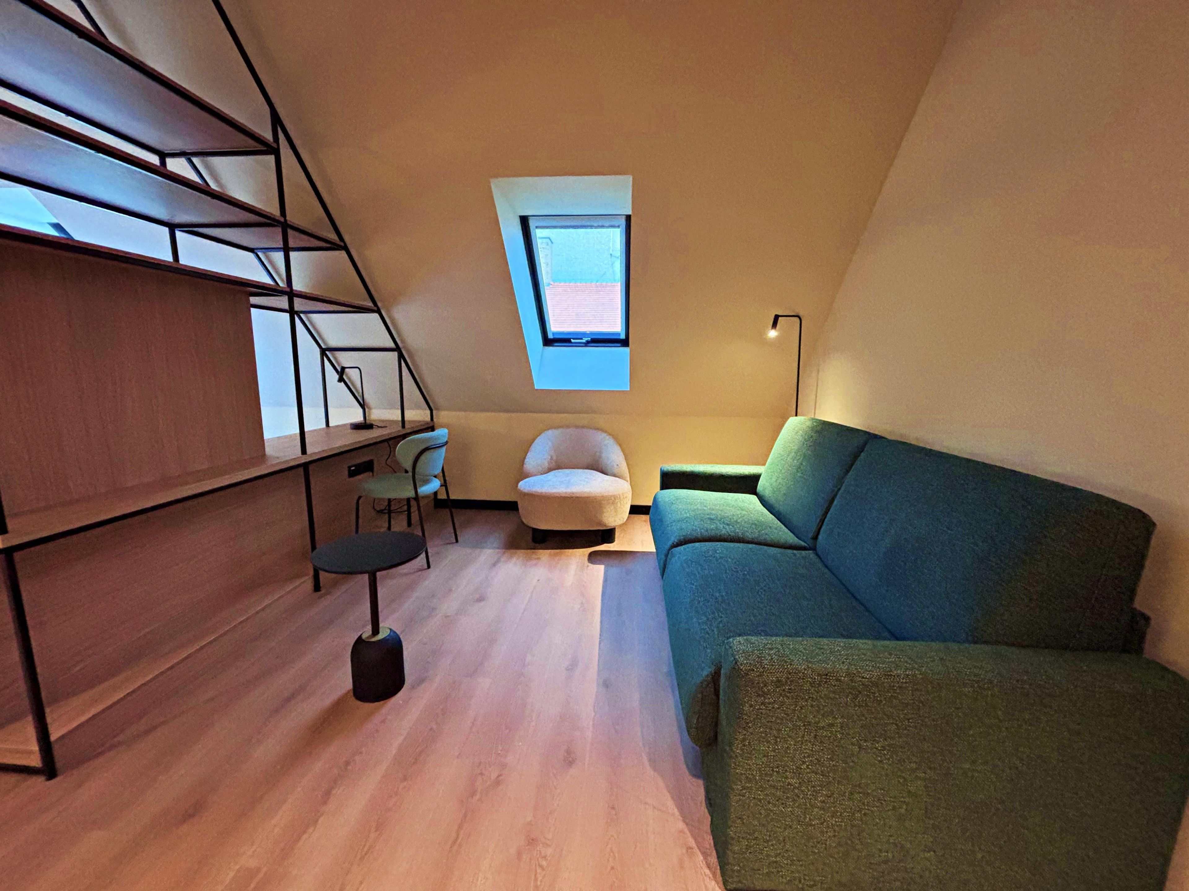 A compact room with a sloped ceiling, a small window, a sofa, a chair, and a desk with a lamp.
