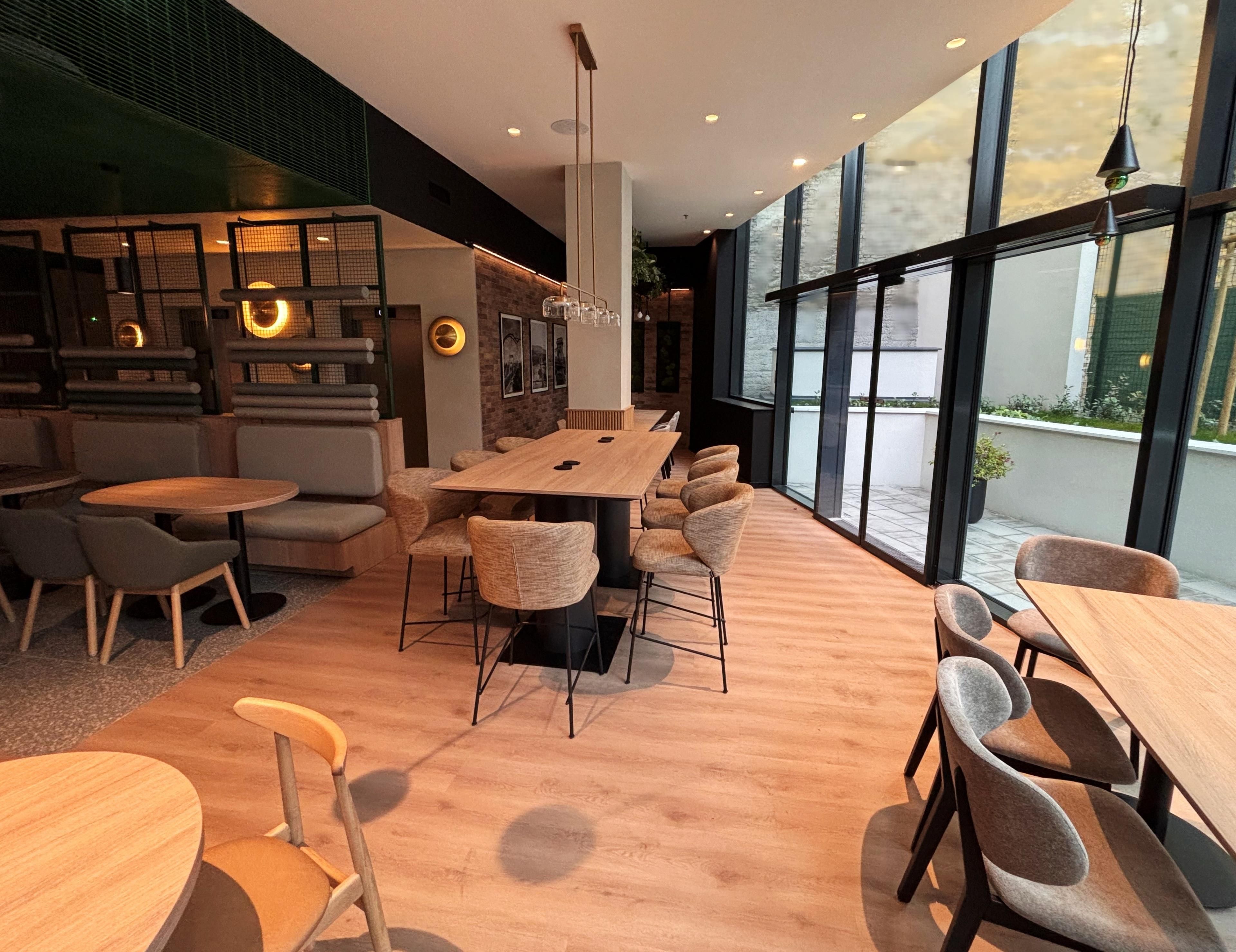 Un interior moderno con una mesa de comedor de madera rodeada de sillas, una zona de bar con taburetes y grandes ventanales que ofrecen luz natural.