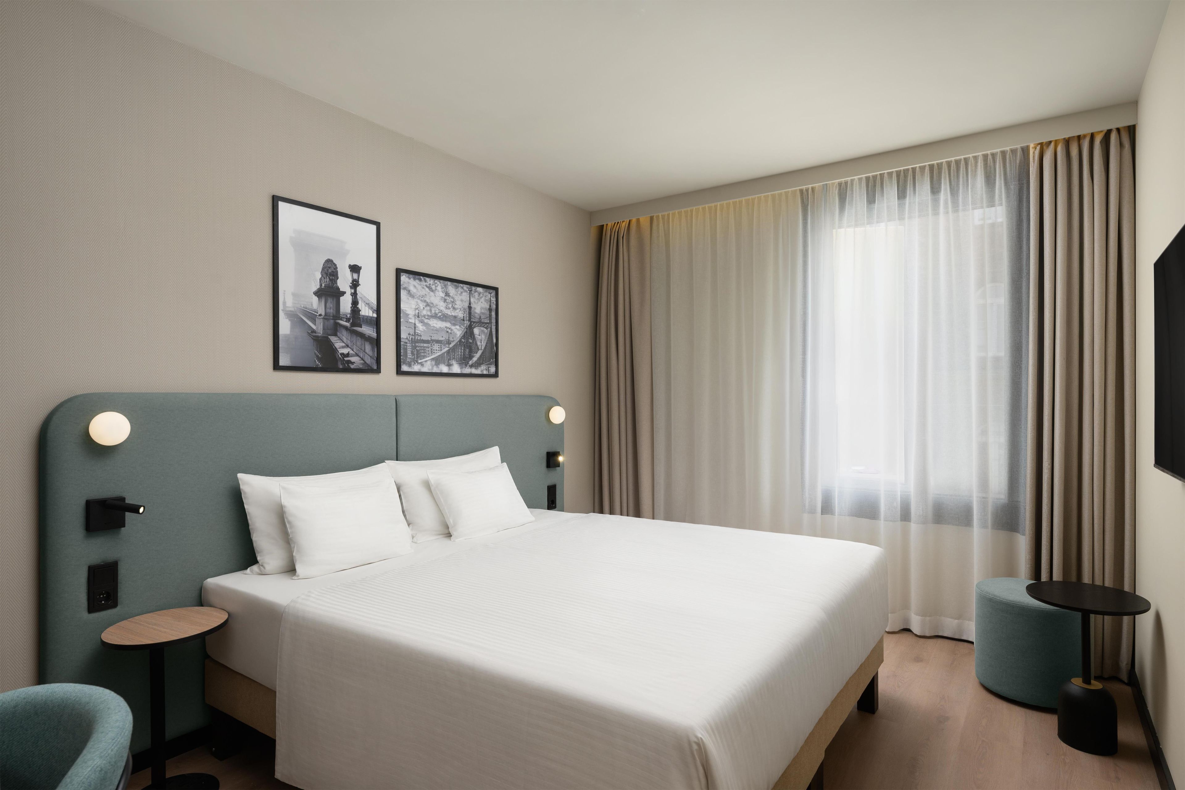 A hotel room with a large bed, two wall-mounted picture frames, a bedside table with a lamp, a round side table, and a large window with curtains.