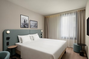 Habitación de hotel con una cama grande, dos cuadros colgados en la pared, una mesita de noche con lámpara, una mesita redonda y un ventanal con cortinas.
