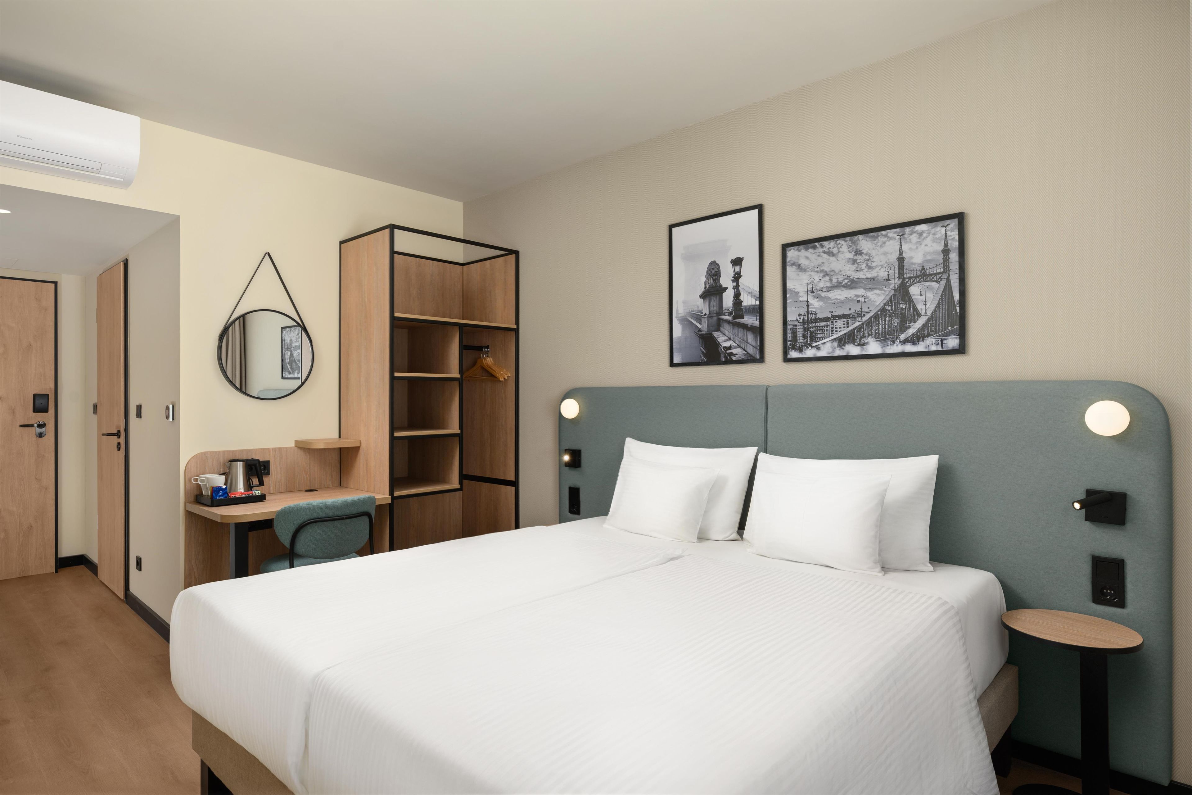 A hotel room with a large bed, a desk, a chair, a wardrobe, and two framed pictures on the wall.