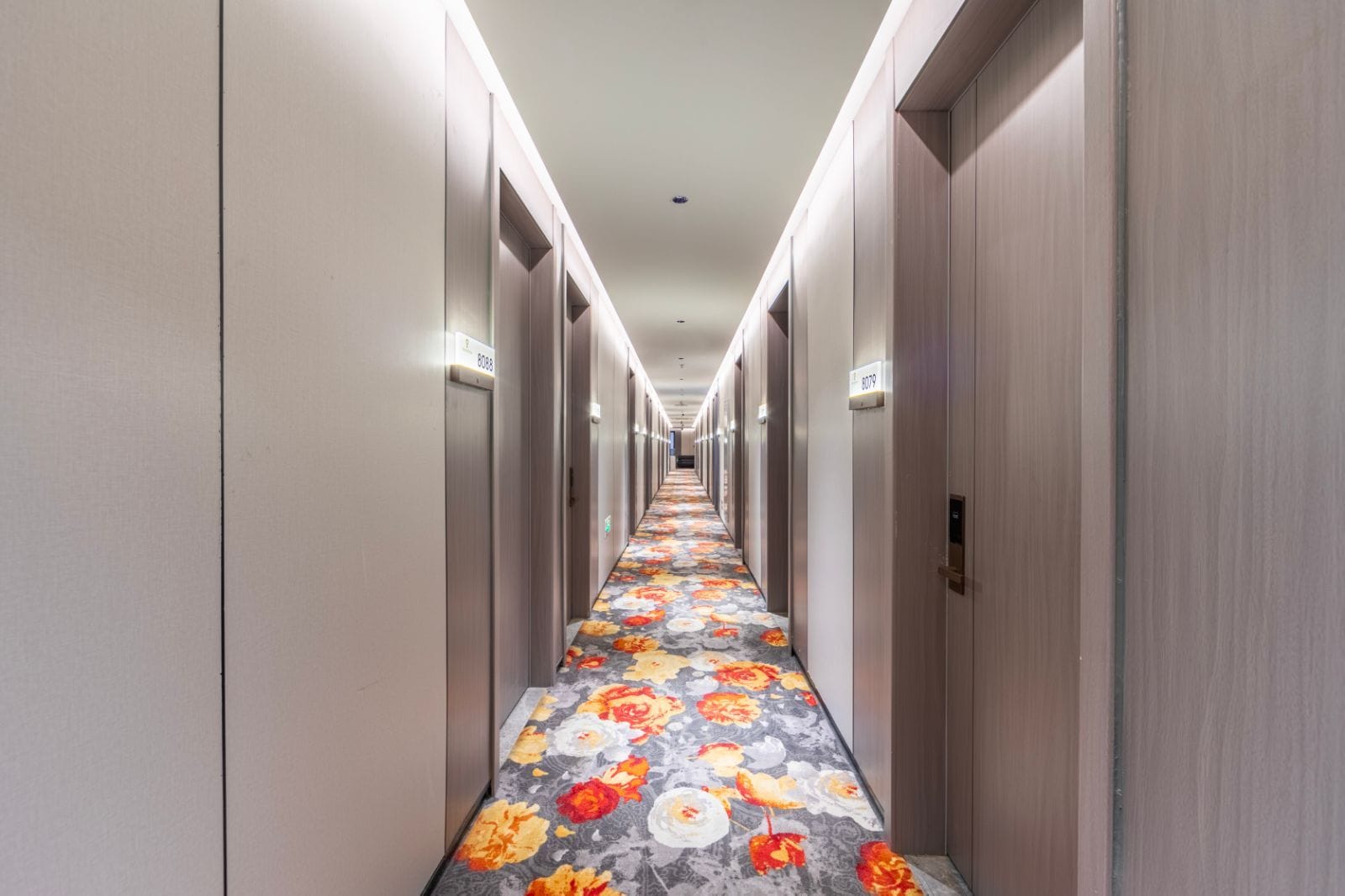 Pasillo de hotel con alfombra estampada con motivos florales, puertas a ambos lados y un cartel con los números de las habitaciones.