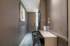 A narrow hallway with a wooden floor, a desk with a chair, a coffee maker, and two water bottles.
