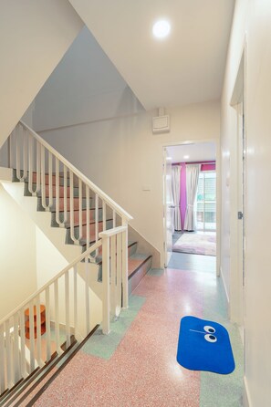 Eine Treppe mit weißen Geländern führt zu einer Zwischenebene, auf dem Boden liegt eine blaue Matte und eine Tür öffnet sich zu einem Raum mit rosafarbenen Wänden und einem Fenster mit Vorhängen.