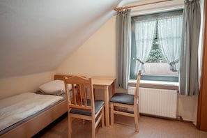 Ein kleiner, dreieckiger Raum mit einem Einzelbett, einem Holztisch und einem Stuhl. Ein Fenster mit Vorhängen bietet Blick auf Bäume.