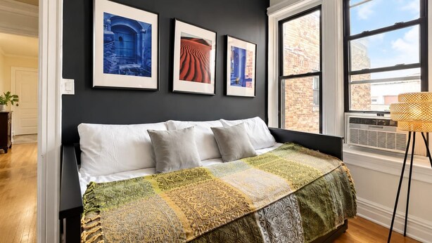 Une chambre à coucher avec un lit, une fenêtre donnant sur des immeubles et trois tableaux encadrés accrochés au mur.