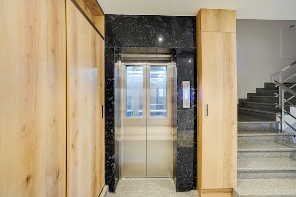 An elevator with a glass door, a wooden wall, and a staircase with metal railings. - Super Townhouse Oak Banaras Central (Varanasi)