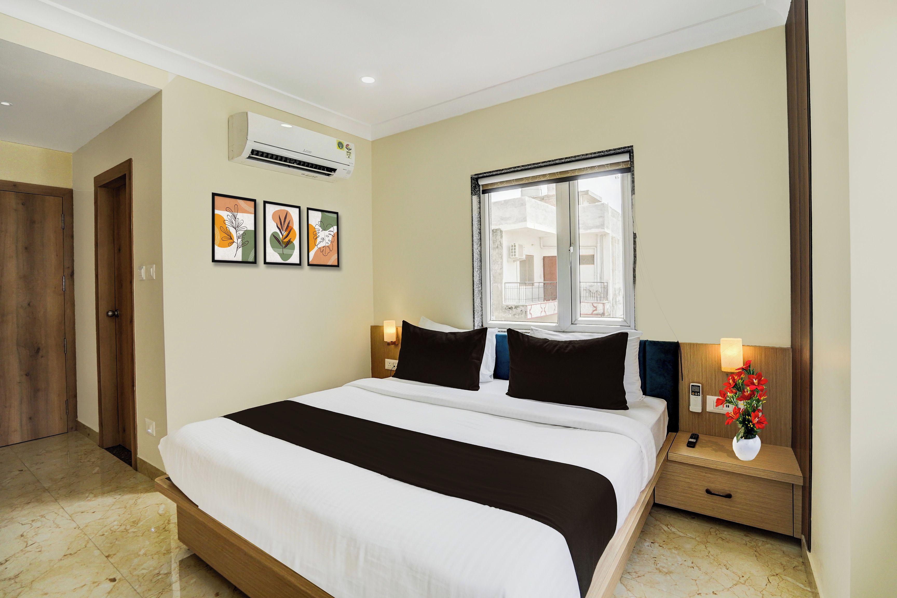 A hotel room with a bed, a window, a nightstand, a wall-mounted air conditioner, and framed artwork.
