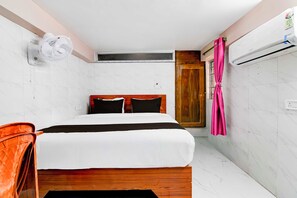 Una habitación de hotel con una cama, un cabecero de madera, una pared blanca, una puerta de madera, una cortina rosa y un equipo de aire acondicionado.