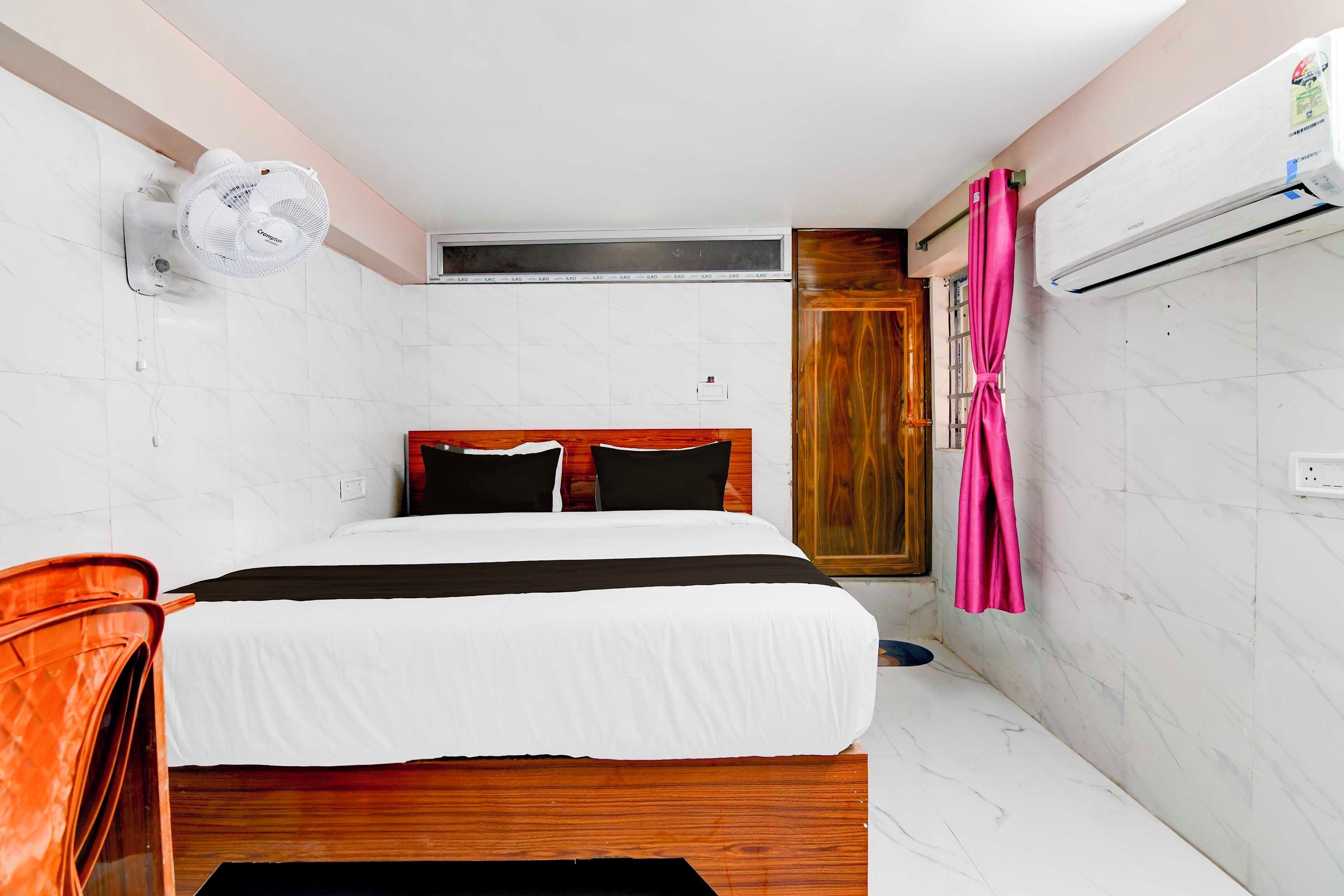 A hotel room with a bed, a wooden headboard, a white wall, a wooden door, and an air conditioning unit.