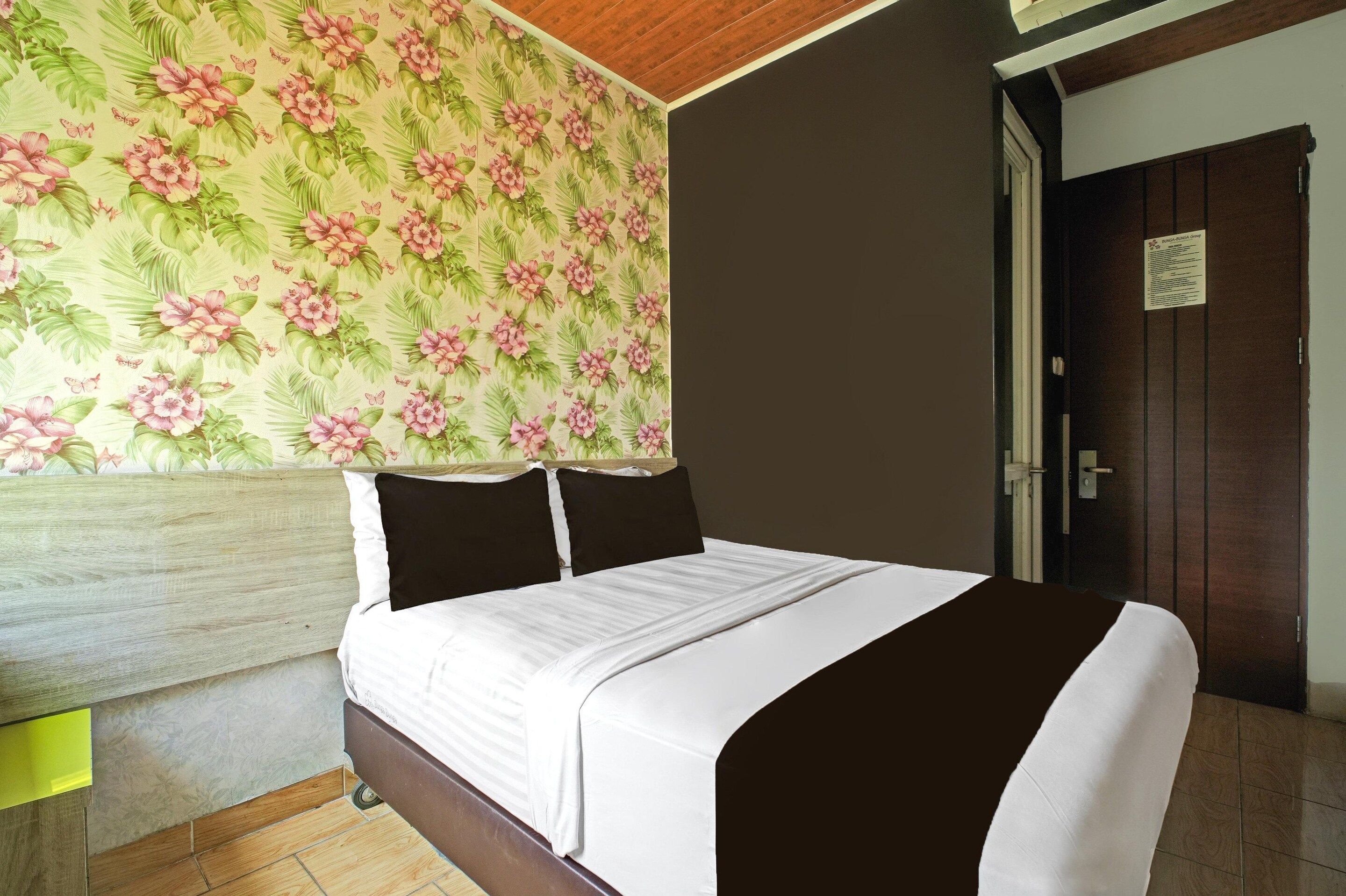A hotel room with a bed, a wooden headboard, a floral wallpaper, a dark door, and a small sign on the door.