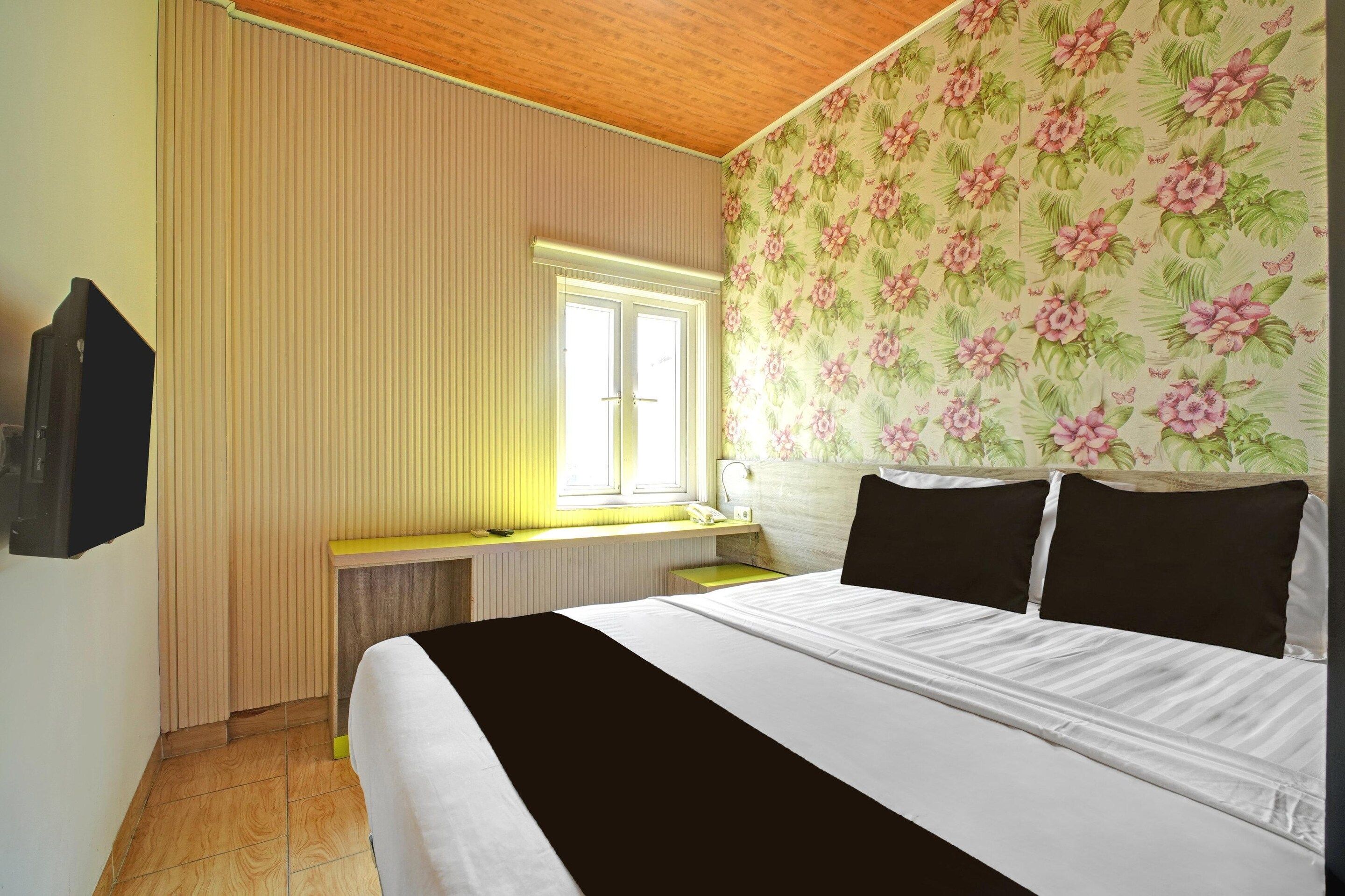A hotel room with a bed, a window with blinds, a desk with a telephone, and a wall with floral wallpaper.