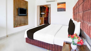 Une chambre d’hôtel avec un lit, un téléviseur à écran plat fixé sur un panneau en bois, un tableau au mur et une plante en pot avec des fleurs.
