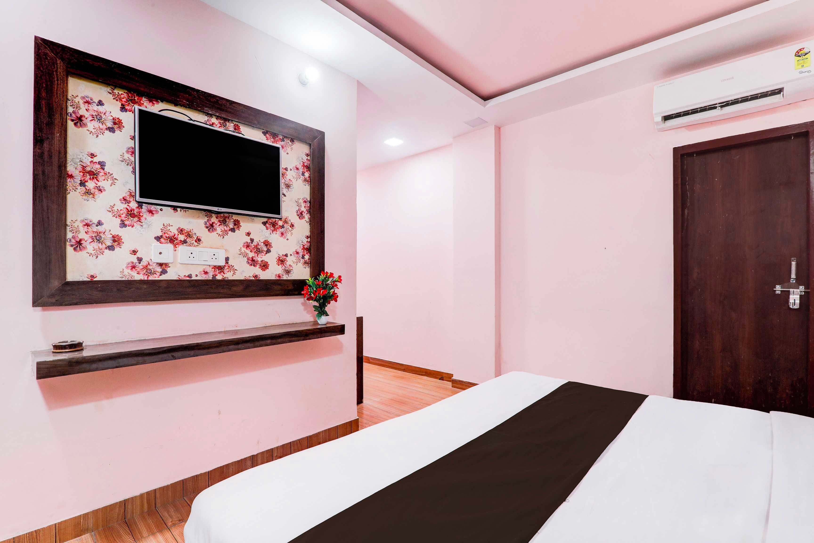Habitación de hotel con cama, televisor montado en una pared con estampado floral, una pequeña estantería con un florero y una puerta de madera.