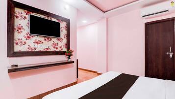 Habitación de hotel con cama, televisor montado en una pared con estampado floral, una pequeña estantería con un florero y una puerta de madera.