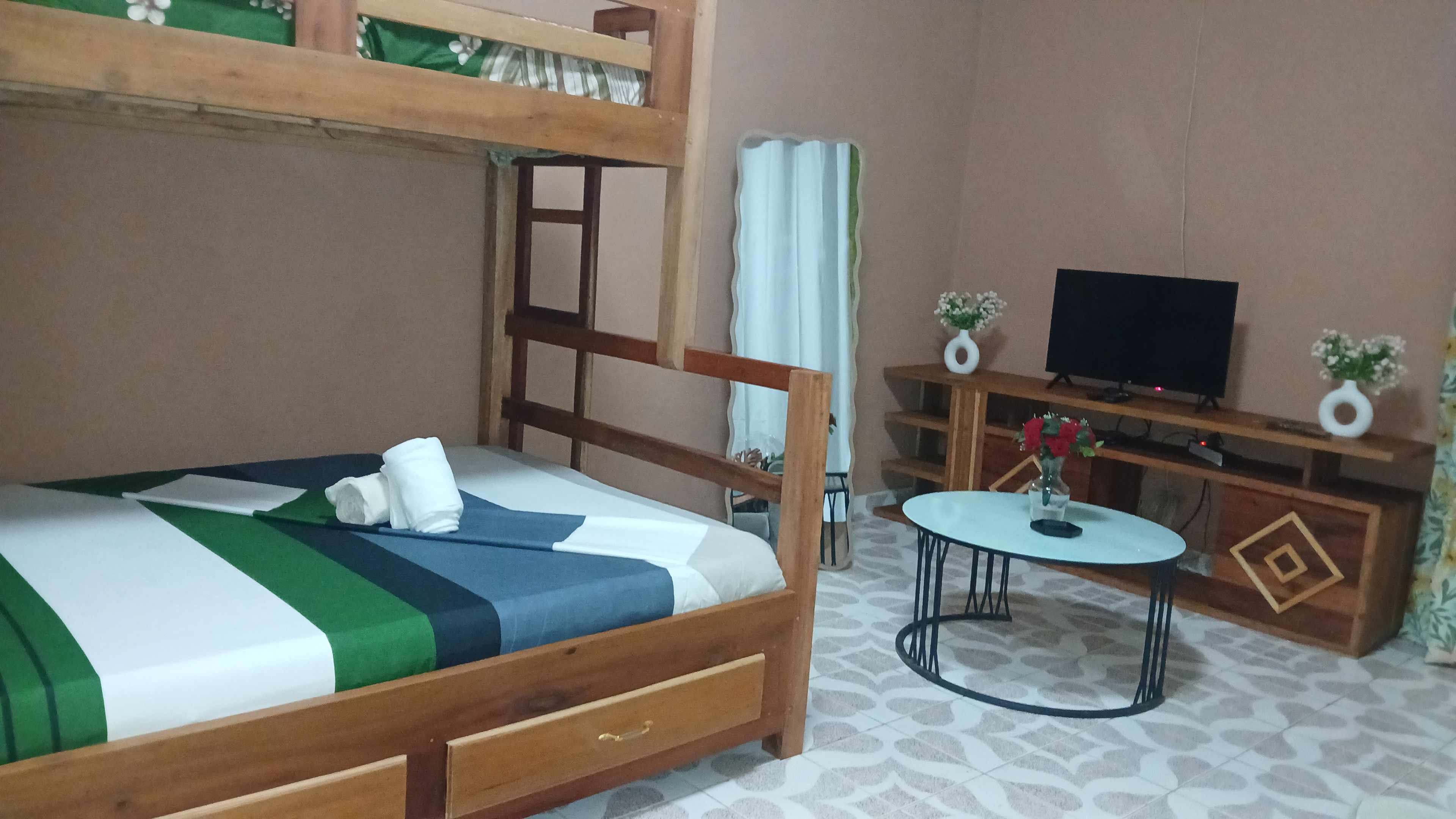 Una cama individual sobre otra con estructura de madera, una mesita redonda con un florero y un televisor sobre un mueble de madera.