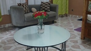 A round glass table with a vase of red flowers in the center, a grey sofa with patterned cushions in the background, and a patterned floor.