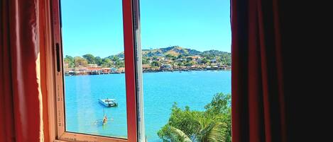 A window with a view of a calm body of water, a boat, and a hilly landscape with buildings.