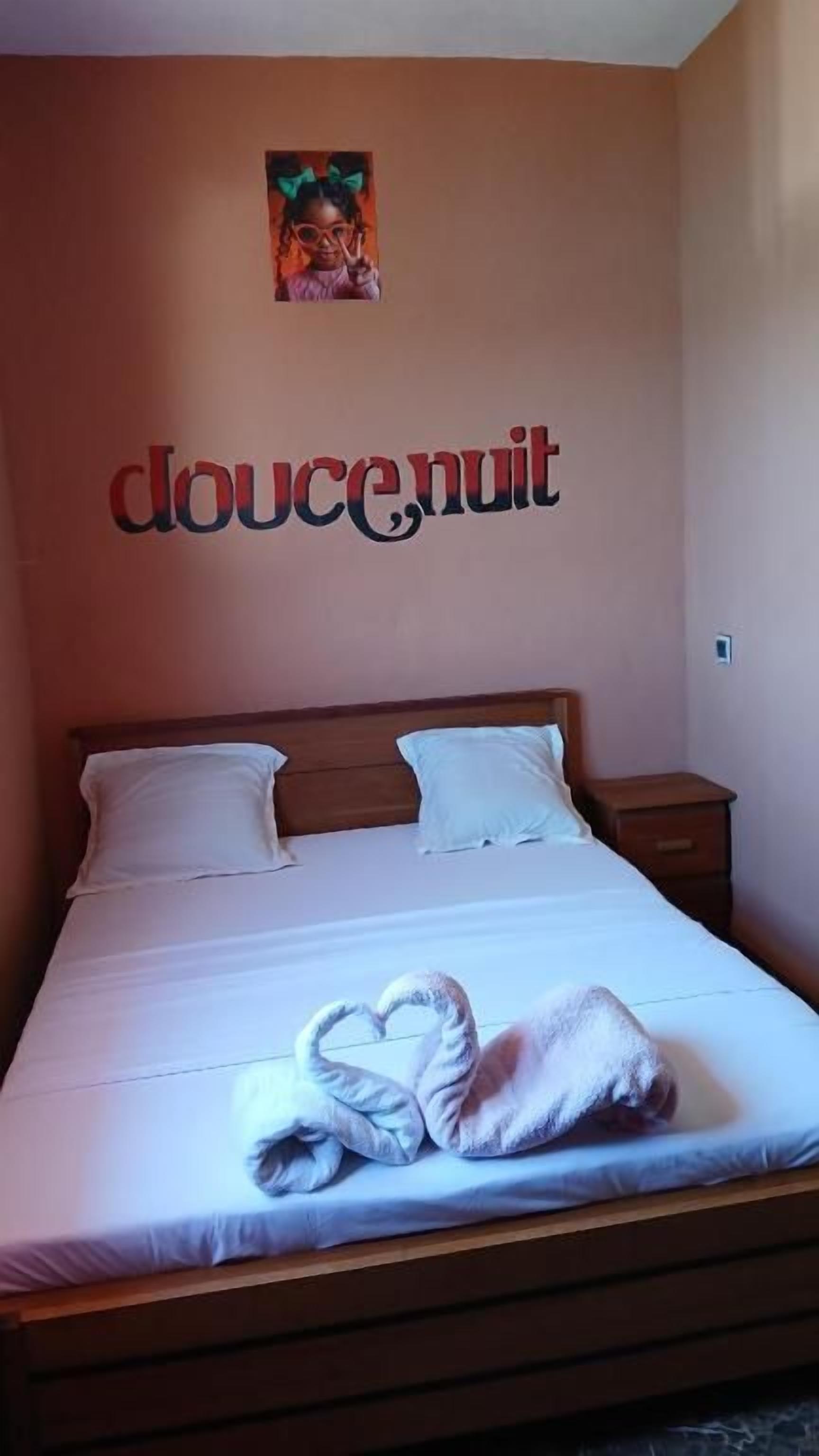 A neatly made bed with a heart-shaped towel and a wall with the word 'doucenuit' in red letters.