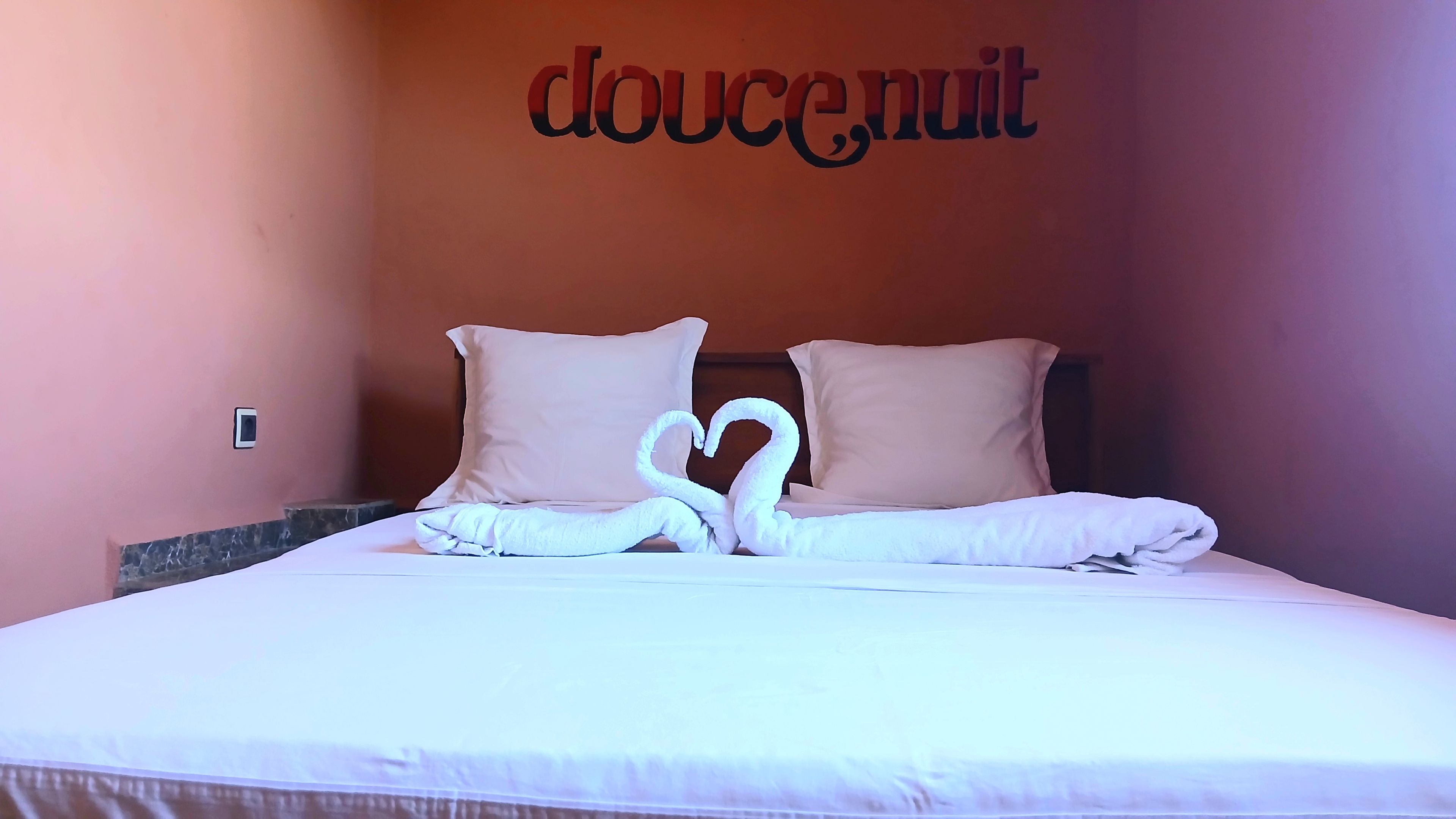 Ein Hotelzimmer mit einem Bett, das eine herzförmige Handtuchanordnung und eine Wand mit dem Schriftzug 'doucenuit' in dekorativer Schrift zeigt.