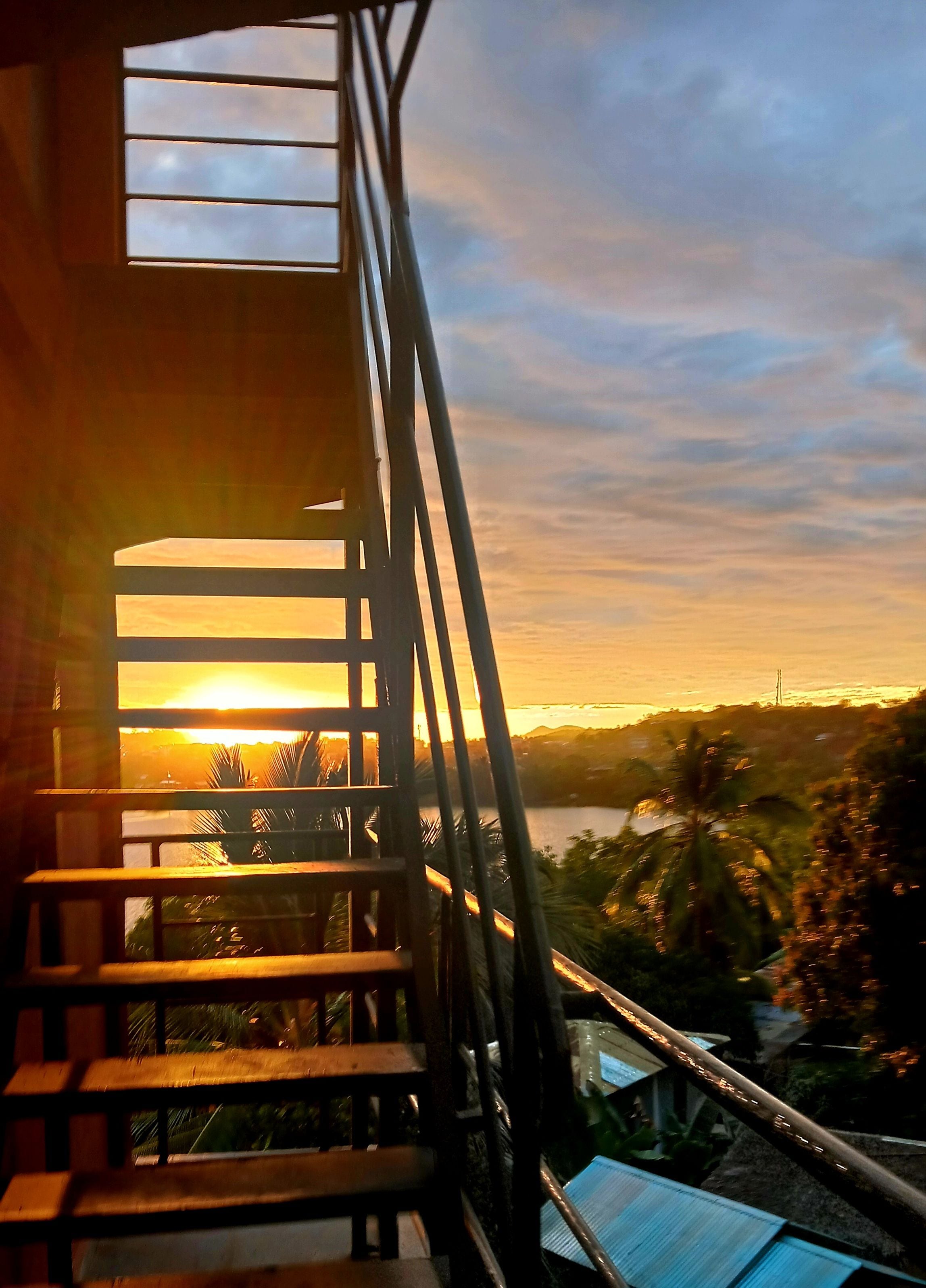 Un escalier menant à un balcon d’où l’on peut admirer un coucher de soleil sur une étendue d’eau.