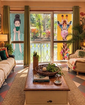 A cozy living room with a sofa, a coffee table with books and a plant, and a view of a garden through a window. - Coco Casita (Woodwark)