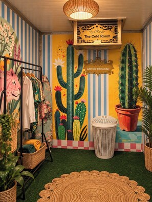 A room with a mural of cacti, a sign reading 'The Cold Room,' and a rack with towels and clothes. - Coco Casita (Woodwark)