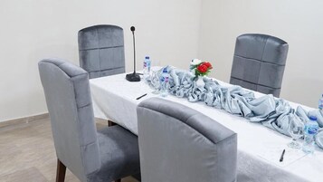 A conference room with a long table, four grey upholstered chairs, a microphone, water bottles, and a red rose on the table.
