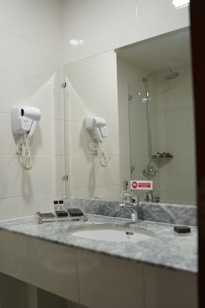 A bathroom with two hair dryers mounted on the wall, a sink with a marble countertop, and a shower area visible in the mirror reflection.