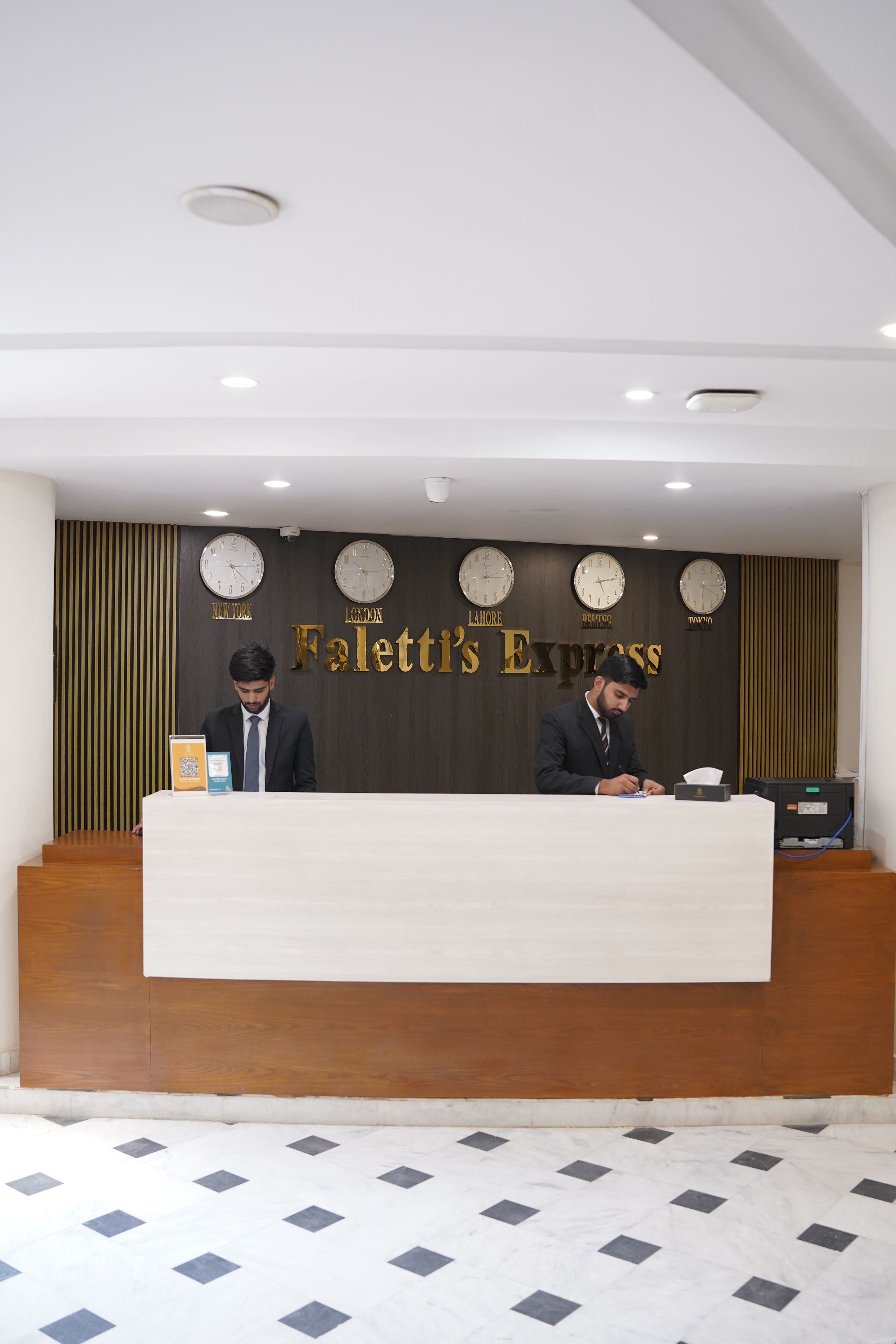 Eine Hotelrezeption mit Uhren, die verschiedene Großstadtdaten anzeigen, einem Schild mit der Aufschrift 'The Falett's Express' und einem modernen Innenraum.