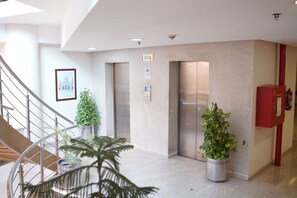 A modern elevator area with a marble floor, two potted plants, a fire hose reel, and a staircase.