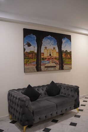 A grey tufted sofa with black cushions against a white wall, with a large painting of a building and landscape above it.