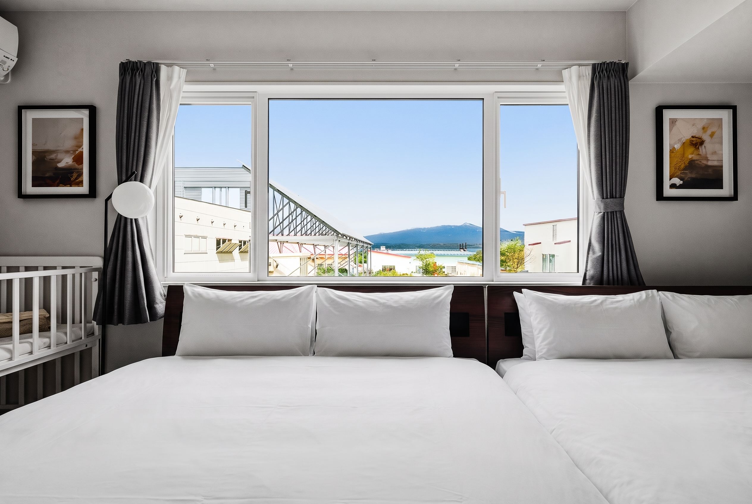 A hotel room with a large bed, a crib, and a view of a building and mountains through a window.