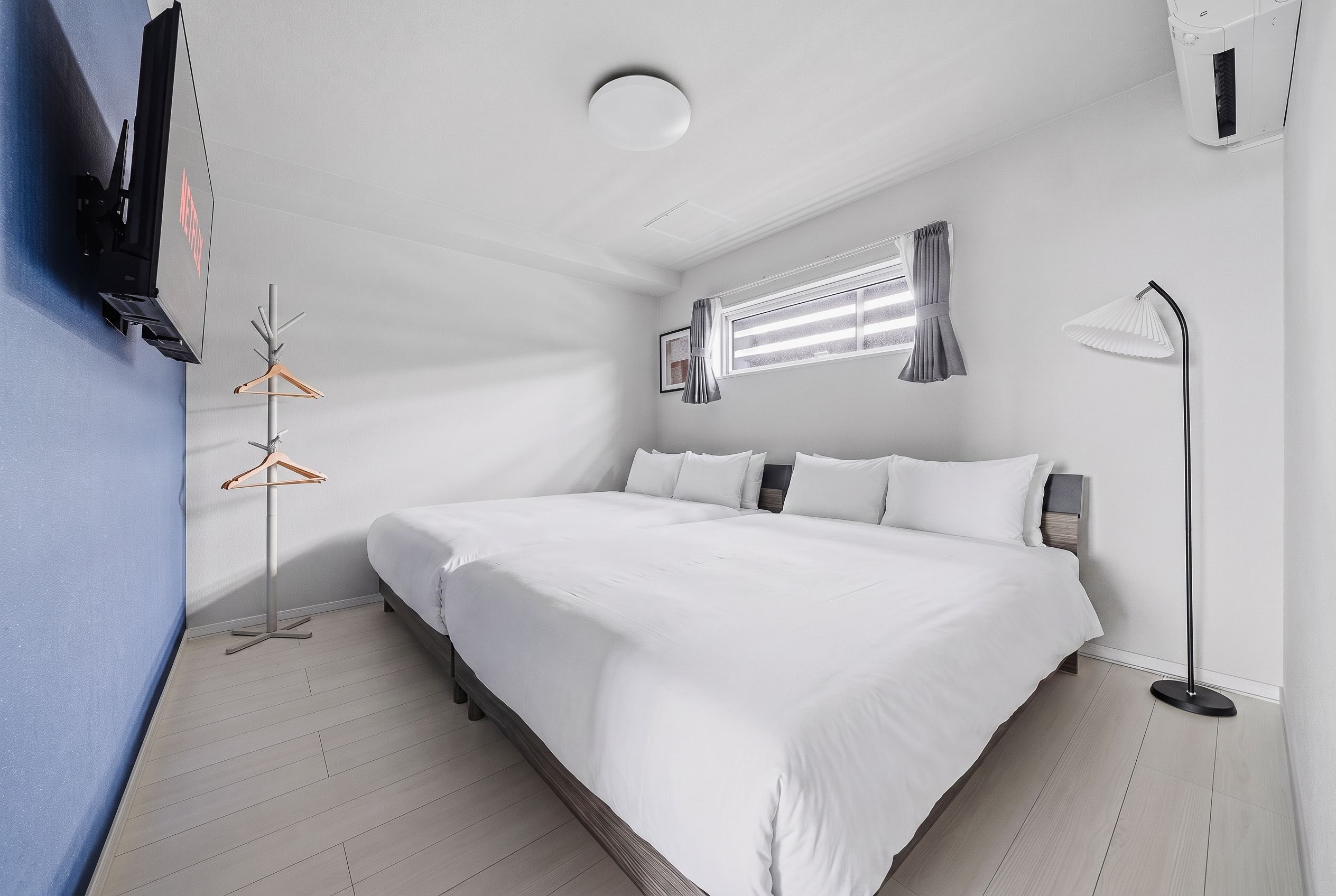 Habitación de hotel moderna con una cama grande, una mesita de noche, un televisor de pared, una ventana con cortinas y una lámpara de pie.