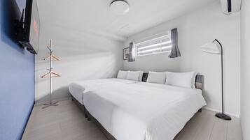 Habitación de hotel moderna con una cama grande, una mesita de noche, un televisor de pared, una ventana con cortinas y una lámpara de pie.