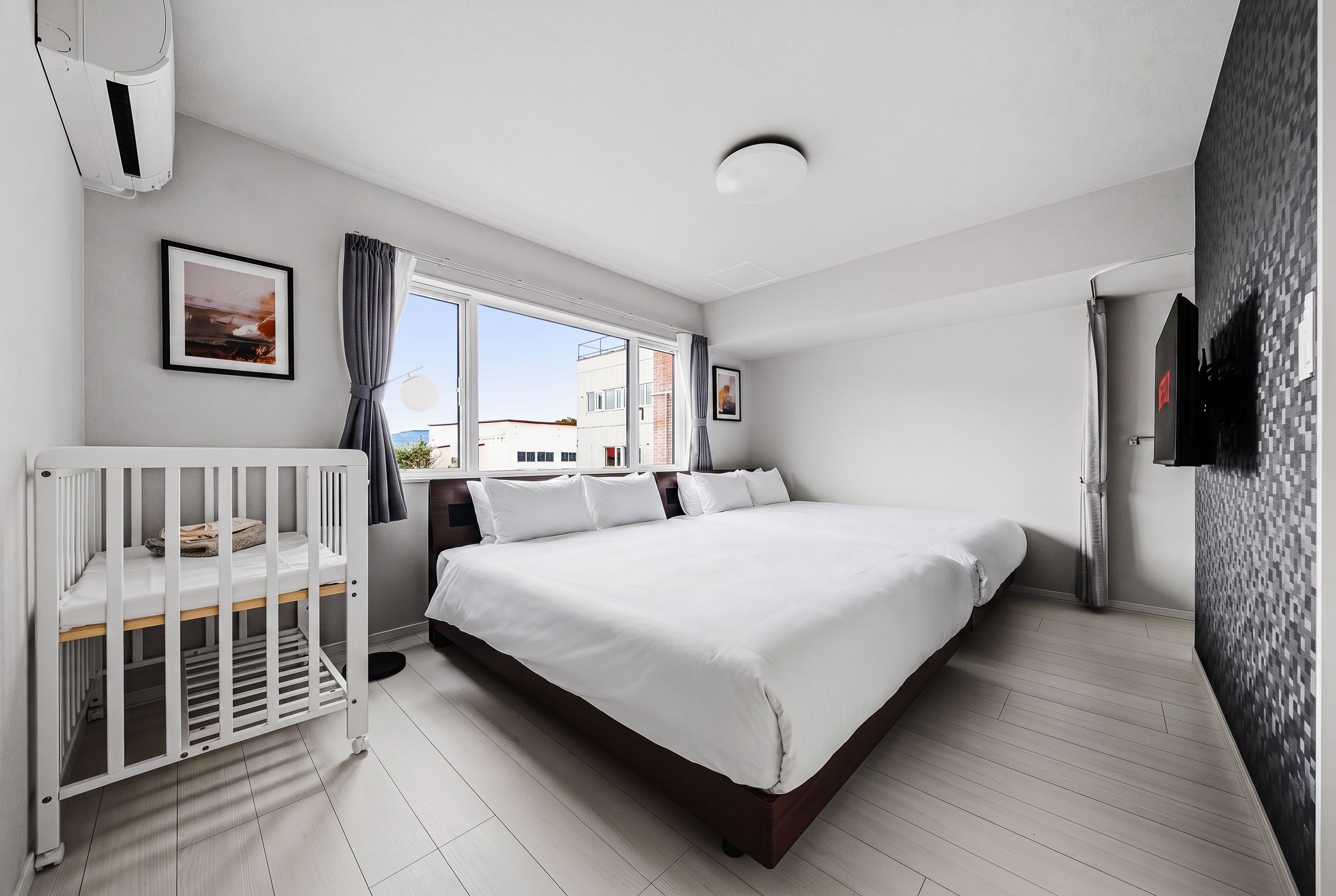 Une chambre avec un berceau, un lit, une fenêtre avec des rideaux et un téléviseur fixé au mur.