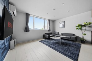 Uma sala de estar moderna com um sof á cinzento, uma mesa de centro, uma televisão e uma janela grande com cortinas.