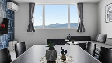 Una habitación de hotel moderna con una mesa de comedor, sillas, un sofá, un televisor y un ventanal con cortinas.