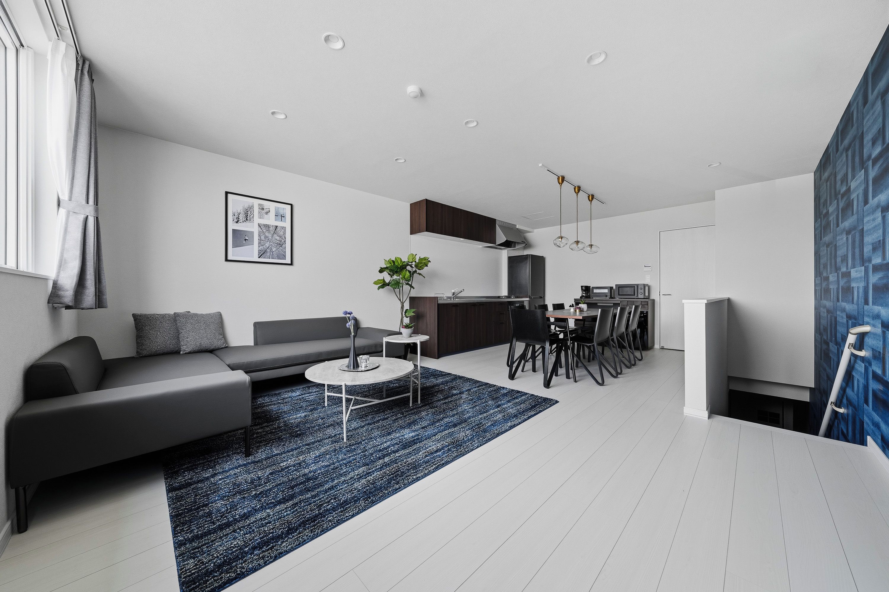 A modern living room with a grey sofa, a coffee table, and a blue rug. There is a kitchen area with a dining table and chairs.