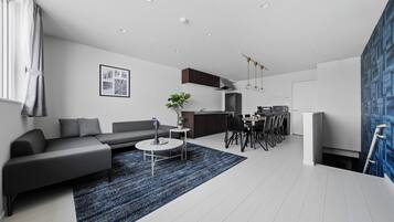 A modern living room with a grey sofa, a coffee table, and a blue rug. There is a kitchen area with a dining table and chairs.