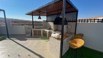 Een terras buiten met een barbecueplek, zitplaatsen en een pergola.
